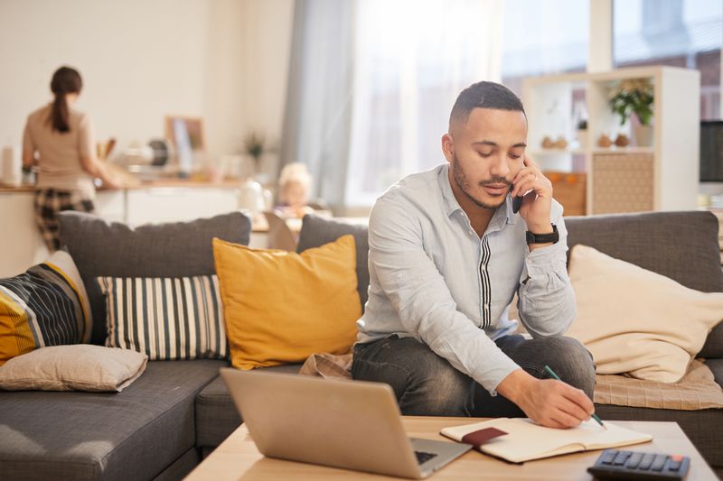 a father working from home on his phone writing on a pad with a laptop in front of him with a wife and child in the back