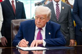 Donald Trump sitting at a desk, signing a document, with others standing in the background