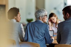 Group of four professionals engaged in a meeting, three facing away and one interacting with the group at a table indoors