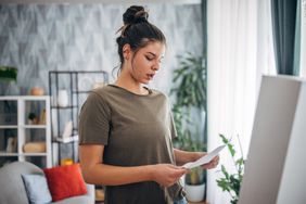 A woman stands in a living room looking concerned as she reads a letter