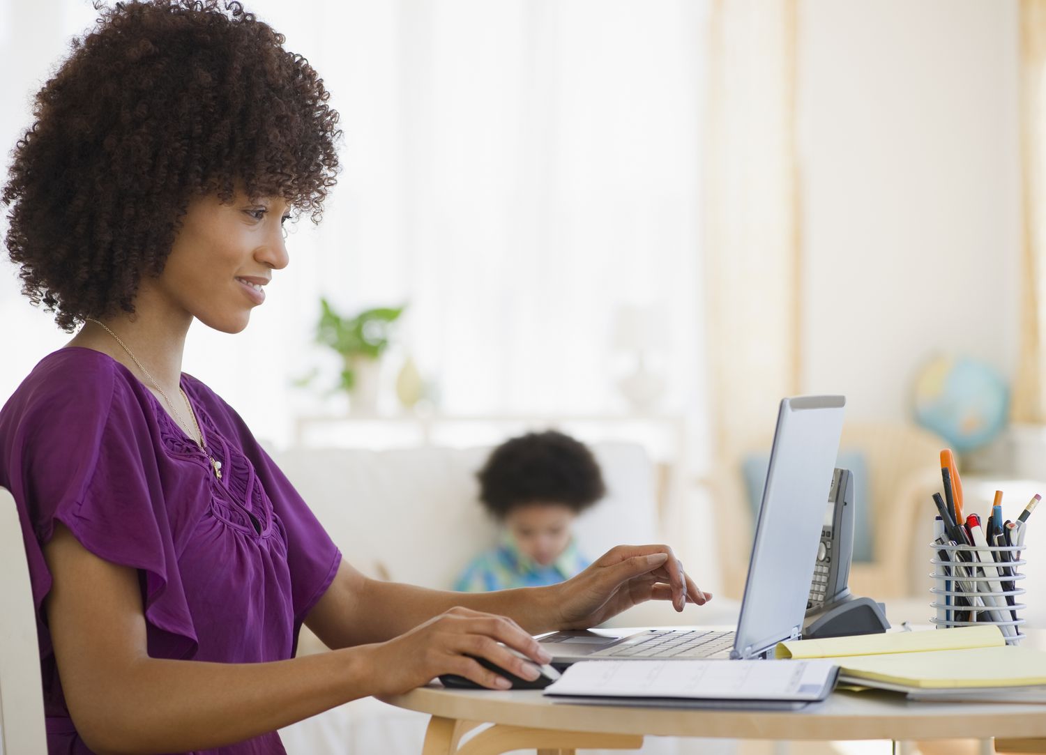 Young mom sitting at a home desk and smiling at something on her laptop, while her young child sits in the background.