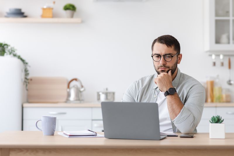 Man in his 30s sitting in his kitchen and pondering something on his laptop