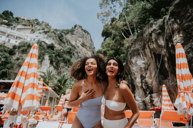 Two women near umbrellas are standing while smiling and laughing in Italy on the beach. 