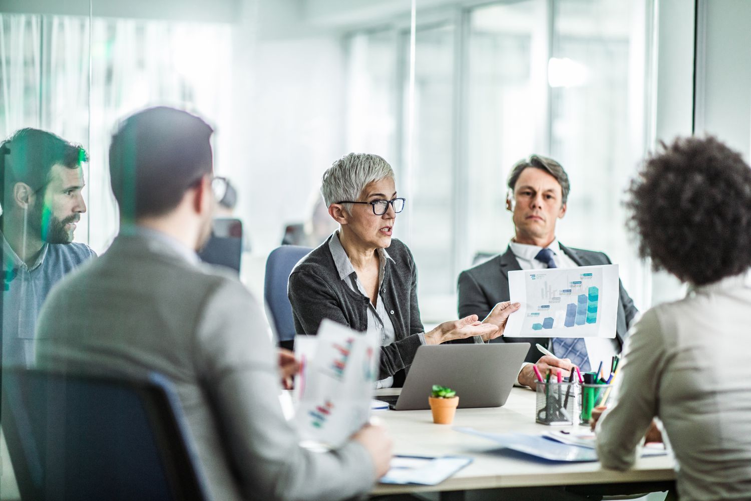 A company board meets around a conference table holding financial documents to discuss financing expansion through the sale of corporate bonds.