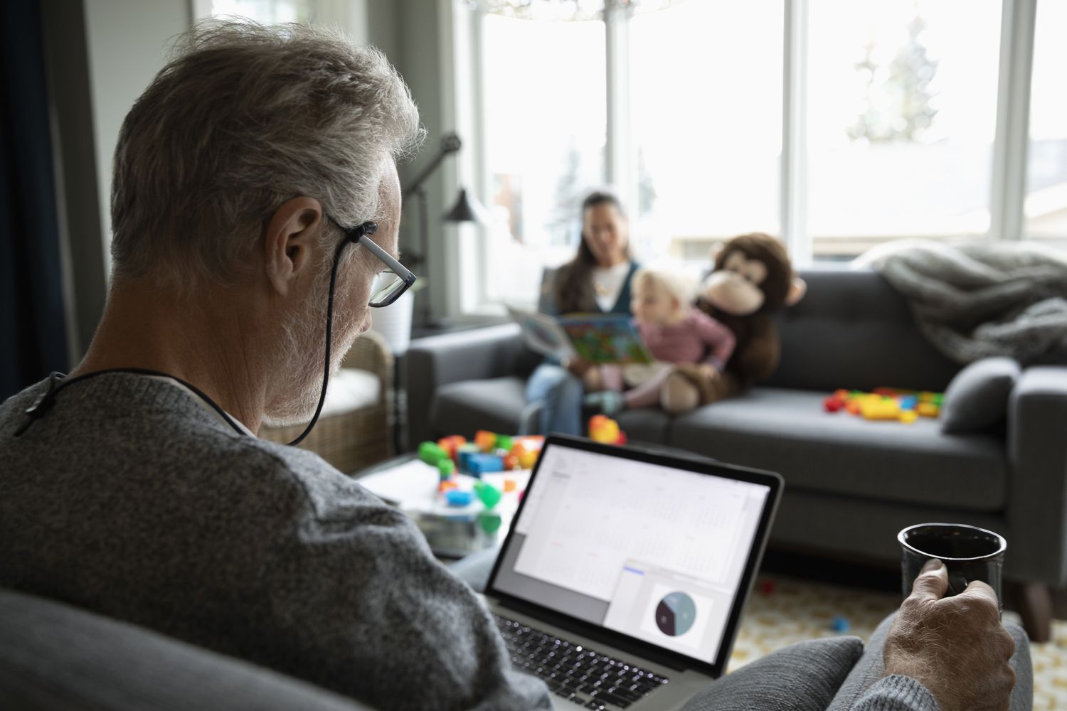 A retiree sits in an overstuffed chair with a mug of coffee reviewing their annuity account on a laptop while their grandchild reads a book with the grandmother on a sofa across the room.