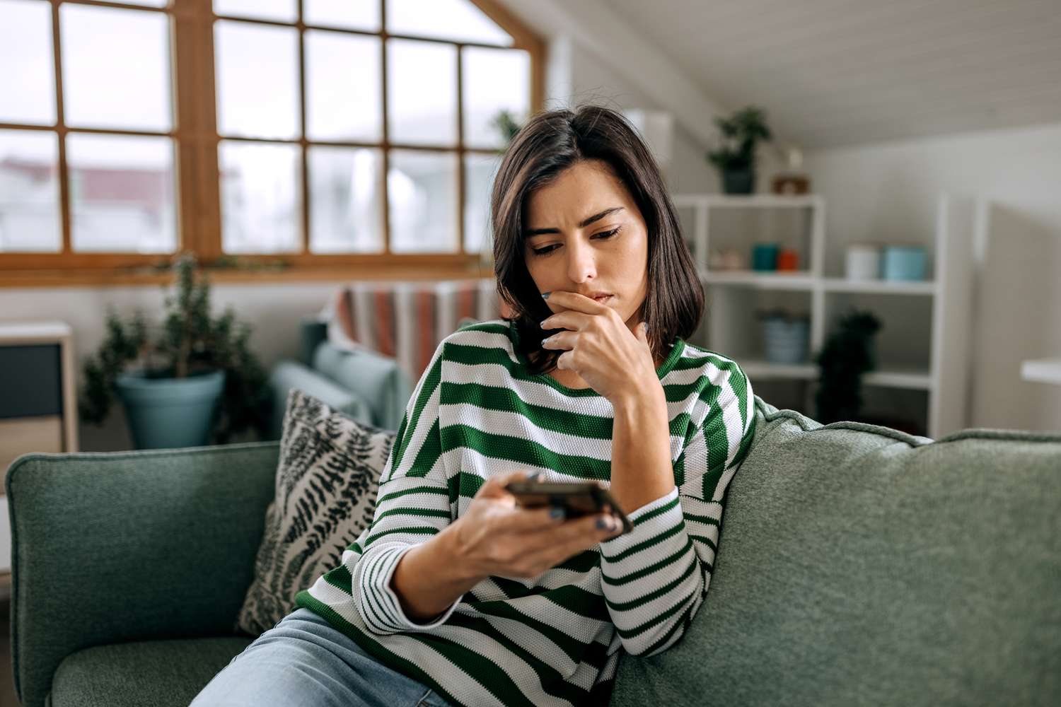 A woman with a worried expression and seated on a couch looks at her smartphone.