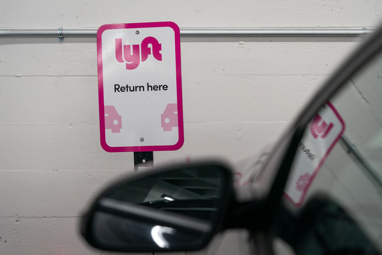 The sign for a Lyft rental vehicle return location is seen between a white wall and a car.