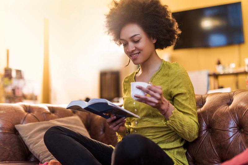 A woman in a green shirt holding a tea cup reads a book on her couch at home