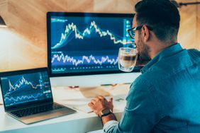 Man sitting at a desk holding a cup of coffee while studying charts on a laptop and monitor.