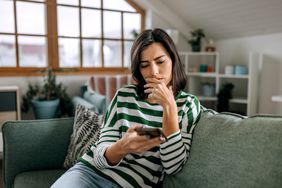 A woman with a worried expression and seated on a couch looks at her smartphone.