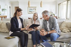 A woman shows a tablet to an older couple seated on a couch, having a conversation in a living room setting