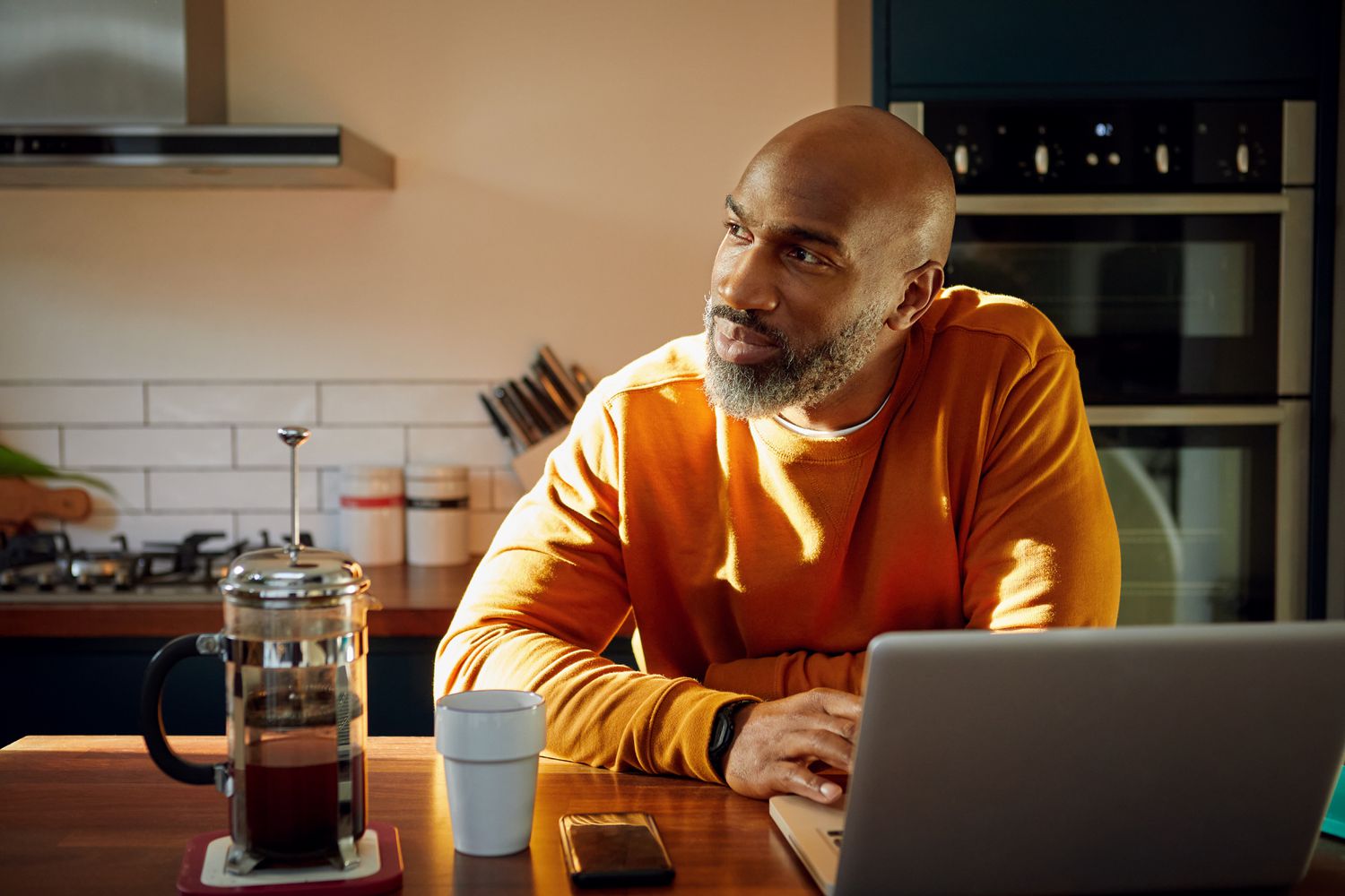 A person sitting at a kitchen table with a laptop and a French press coffee maker