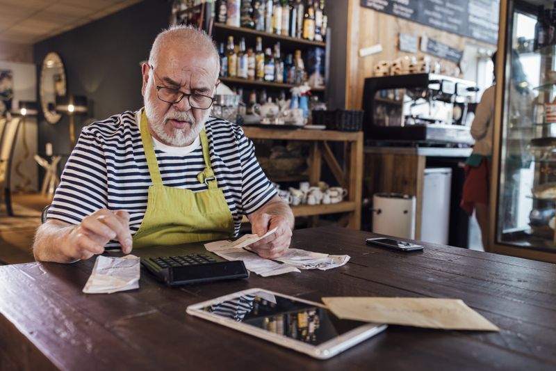 Man wearing an apron while sitting at a table in his cafe adding up receipts on a calculator.