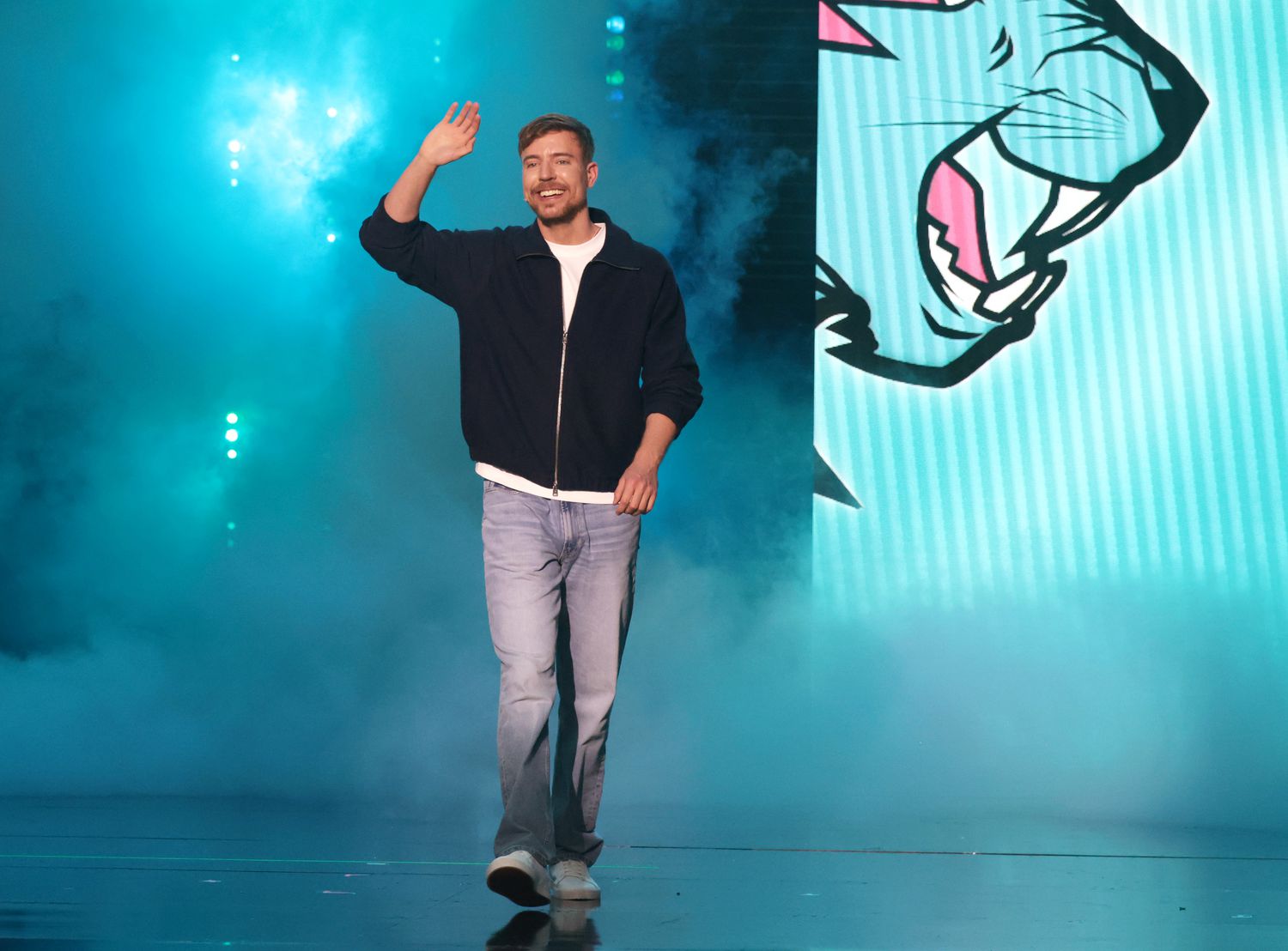 MrBeast waving while walking on stage with blue lighting and a graphic design background
