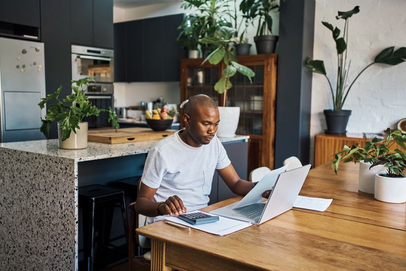 A man sits at a dining room table with a laptop, papers, and a calculator