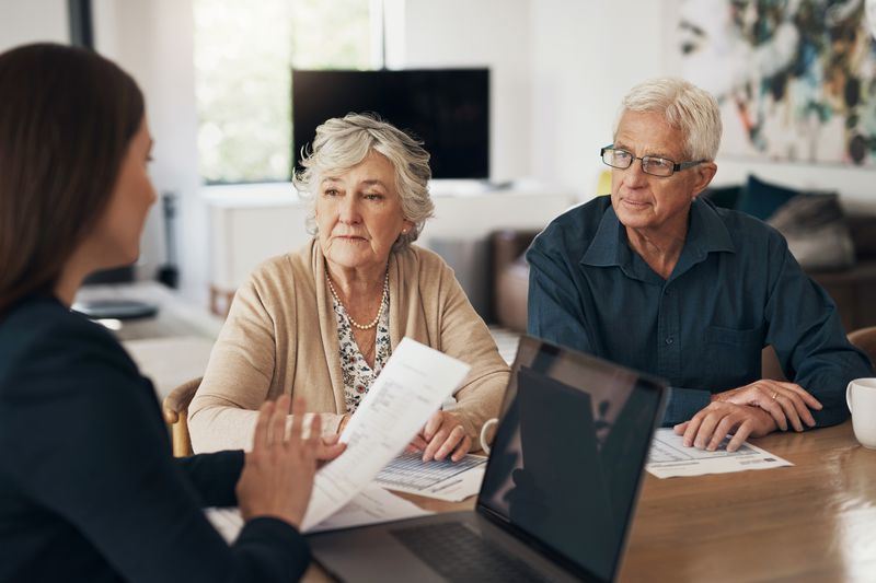 Woman talking to senior couple with paperwork in hand and a laptop in front of her. 