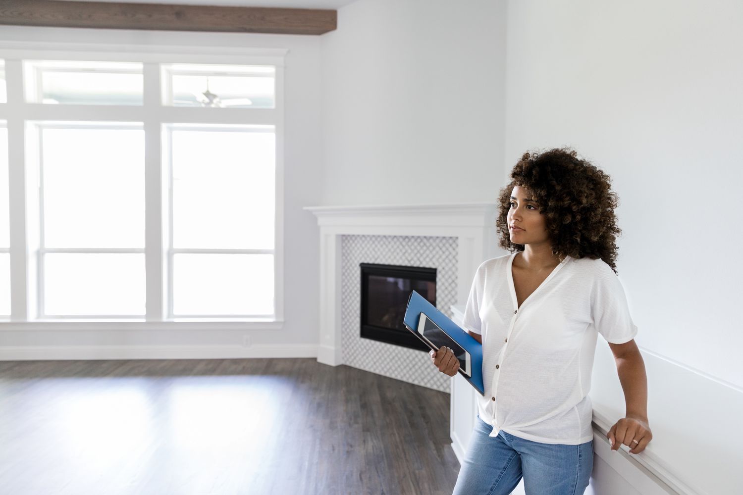 A woman stands in an empty living room holding a folder and tablet