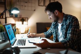 Person writing at a desk with a laptop, phone, and papers in a dimly lit room