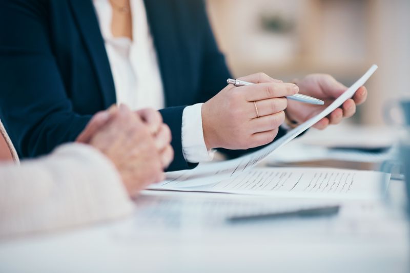 A close up showing the arms and hands of two people at a desk while one of them reads over an insurance policy before signing the document.
