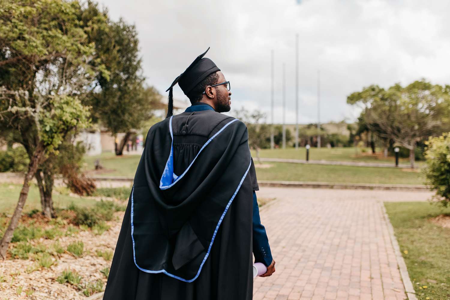 Young male graduate with beard wearing graduation gown and mortarboard walking and looking sideways into distance on campus grounds