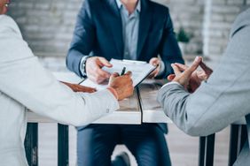Three people sit at a desk, while one person signs a legal document
