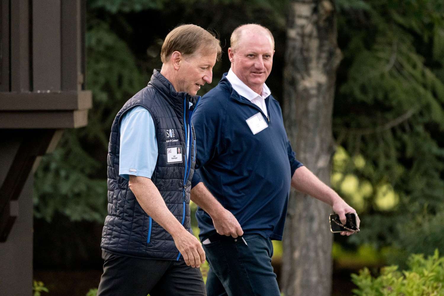 Lee Styslinger and Greg Abel walk next to each other, with trees and bushes in the background.