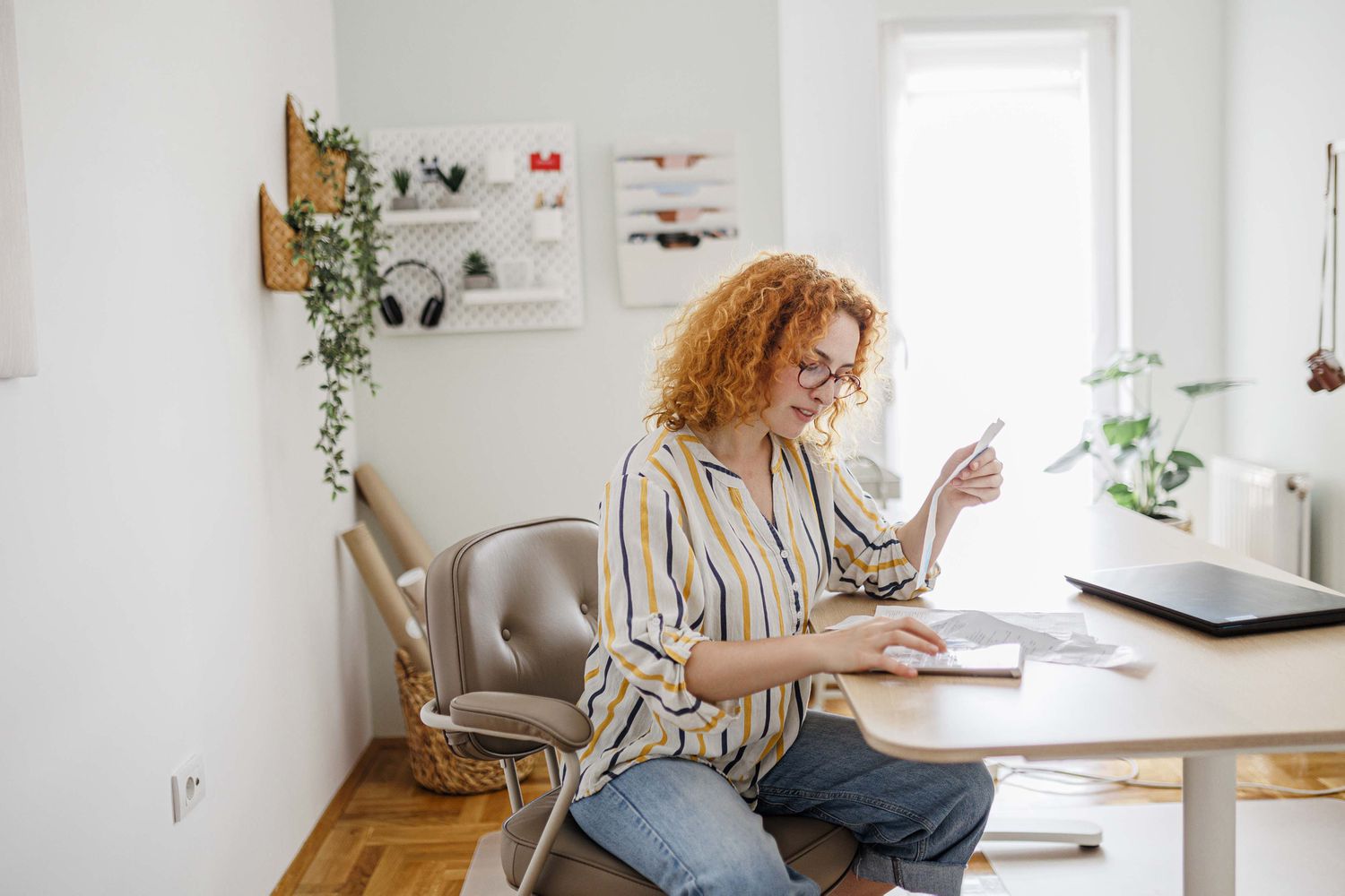 This image shows a person with red curly hair, wearing a striped shirt and jeans, sitting in a comfortable chair at a desk. They are holding papers, possibly reviewing them, with a notebook and a laptop on the desk. The setting is a cozy and well-lit room with modern decor, including plants, wall organizers, and wooden flooring. 