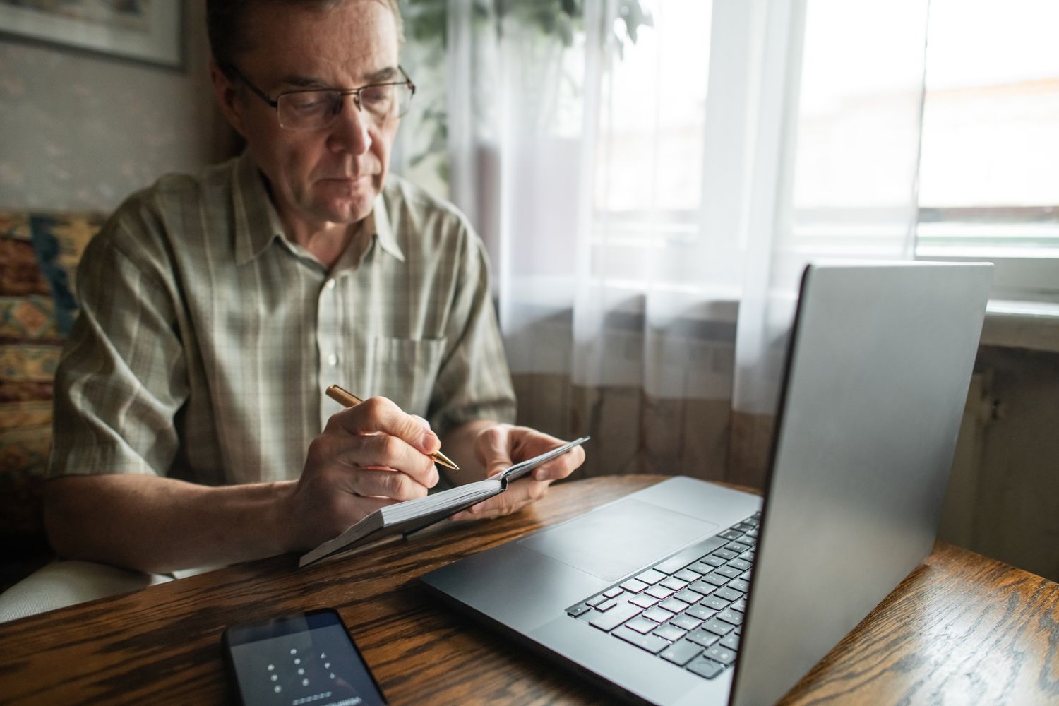 Older man writing notes in a notebook while in front of computer