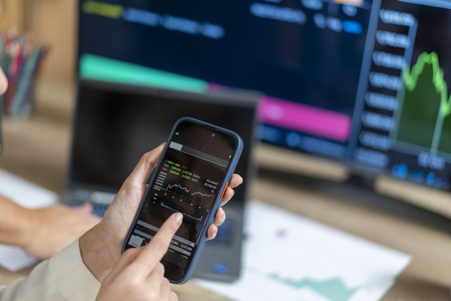 A woman holds a cell phone to check stock market prices, with a computer screen in the background.