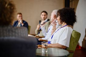Panelists sitting, woman is drinking and two are holding microphones.