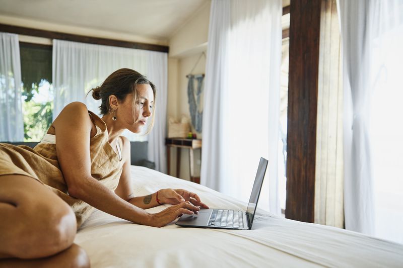 Woman in Overalls Lies on Bed and Looks at Laptop