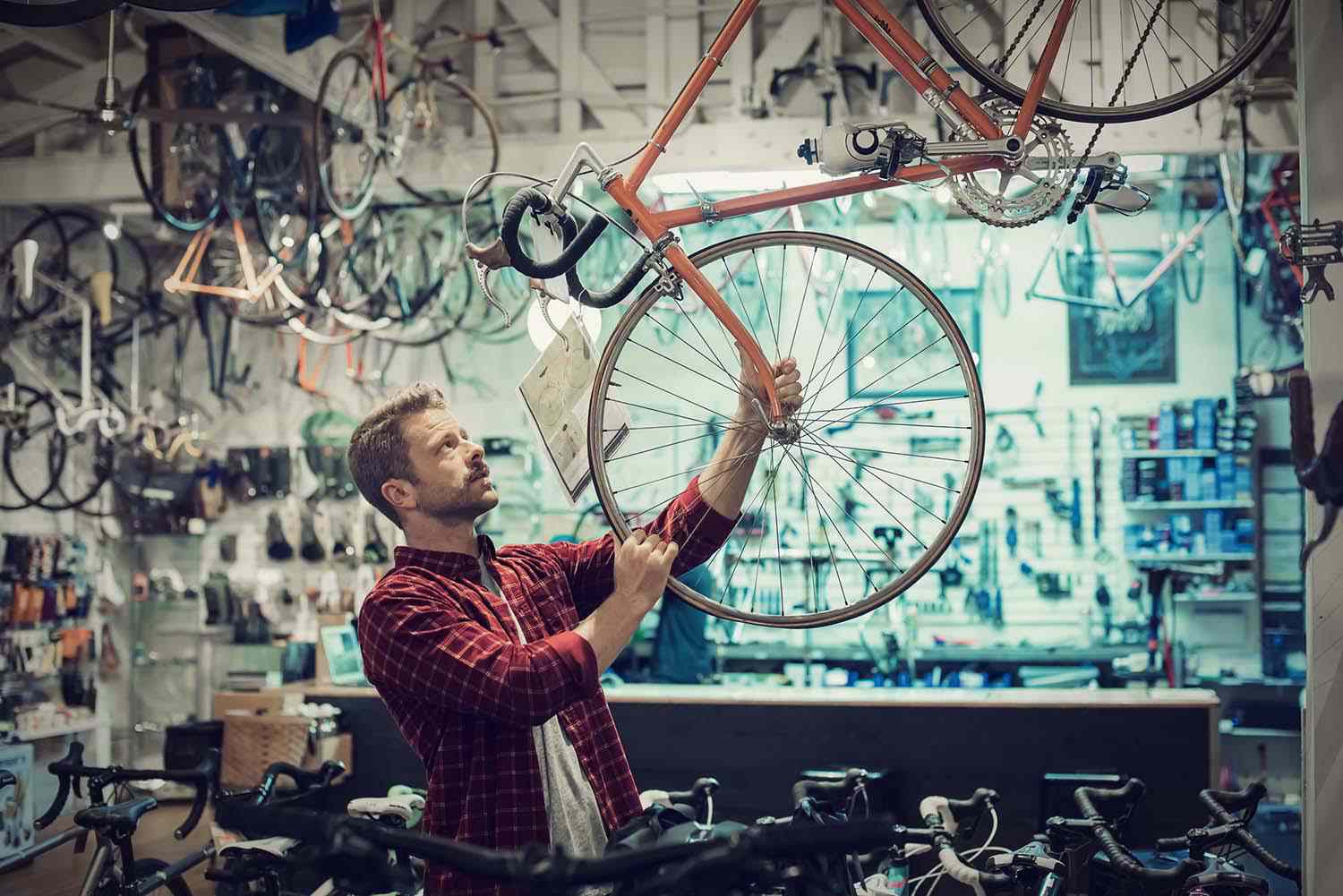 The image shows a man in a plaid red shirt inspecting a bicycle hanging from a display in a bike shop. He appears focused as he examines the wheel or frame. The shop is filled with various bicycles and bike parts hanging from the ceiling and displayed on shelves and racks in the background, creating a busy, well-stocked atmosphere. The scene conveys attention to detail and an interest in cycling.