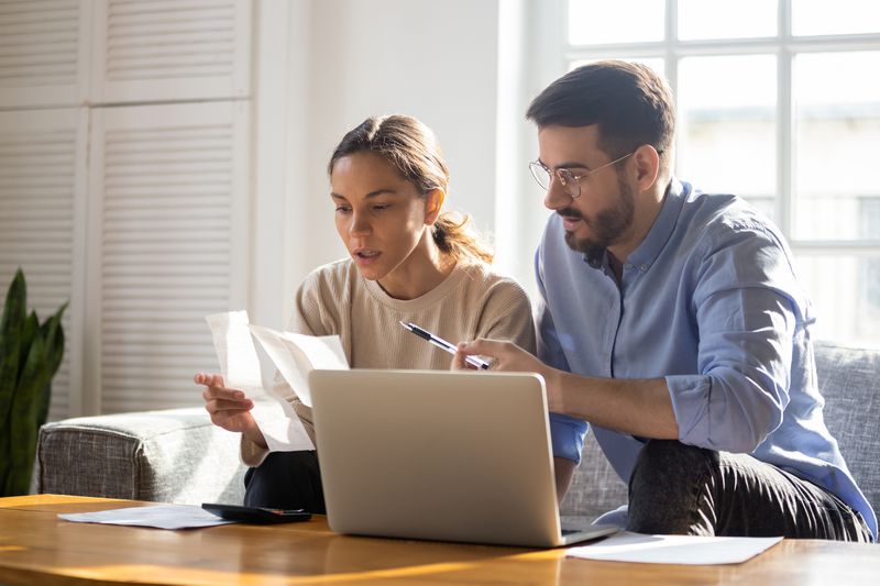 Young couple sitting on their sofa and looking together at financial documents and a laptop