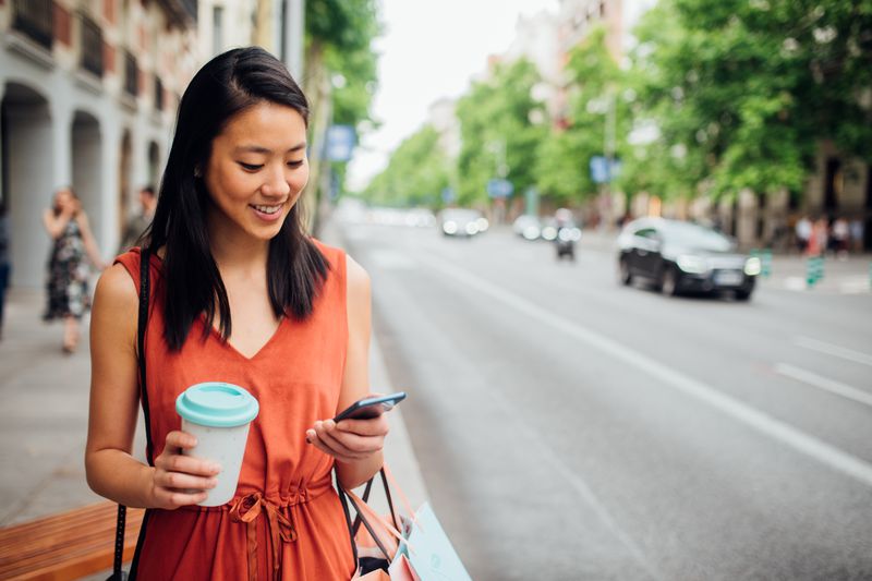 A woman in an orange dress stands on a street with a coffee and smartphone in hand
