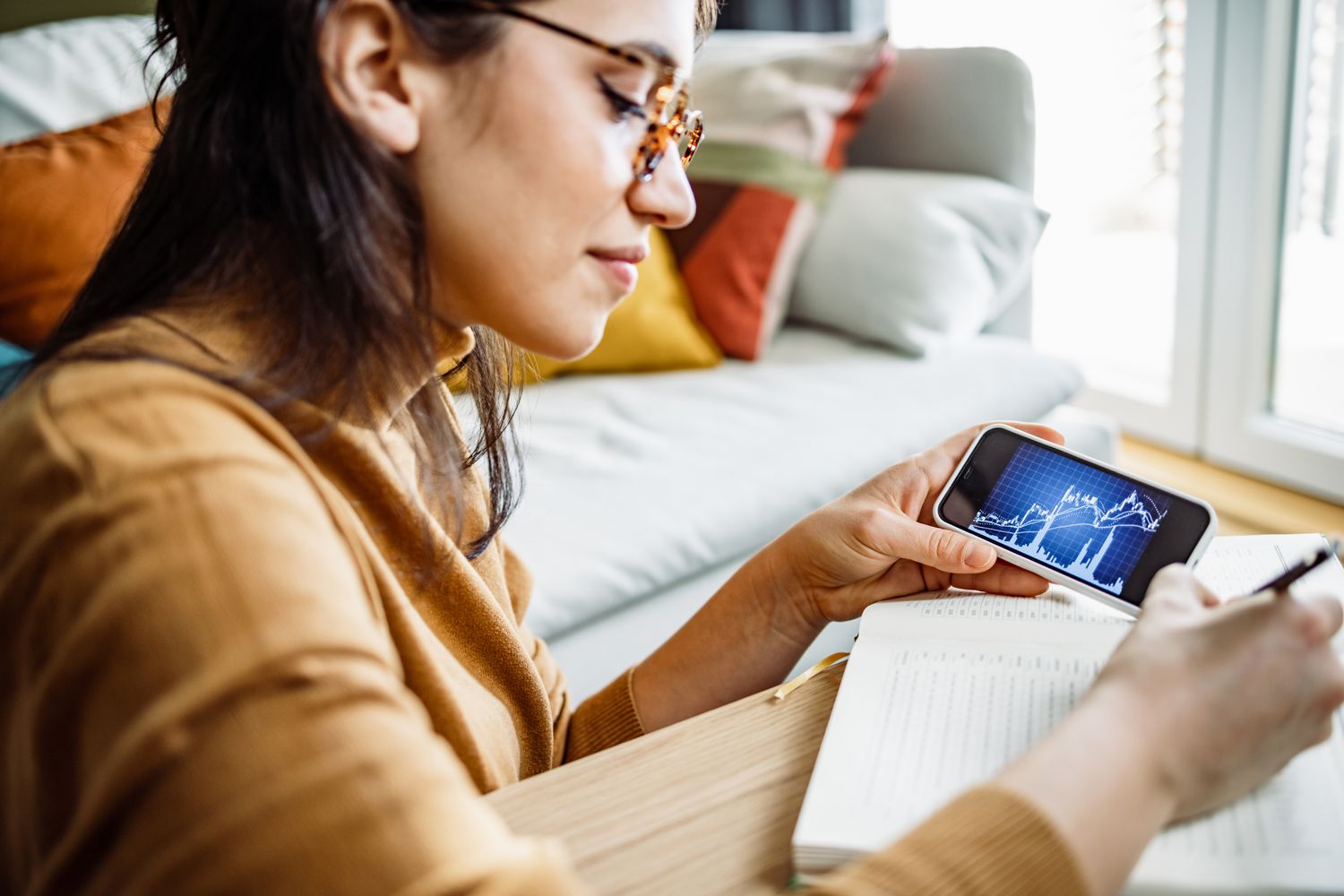 Woman sitting on the floor in her living room looking at a chart on her phone and making notes.