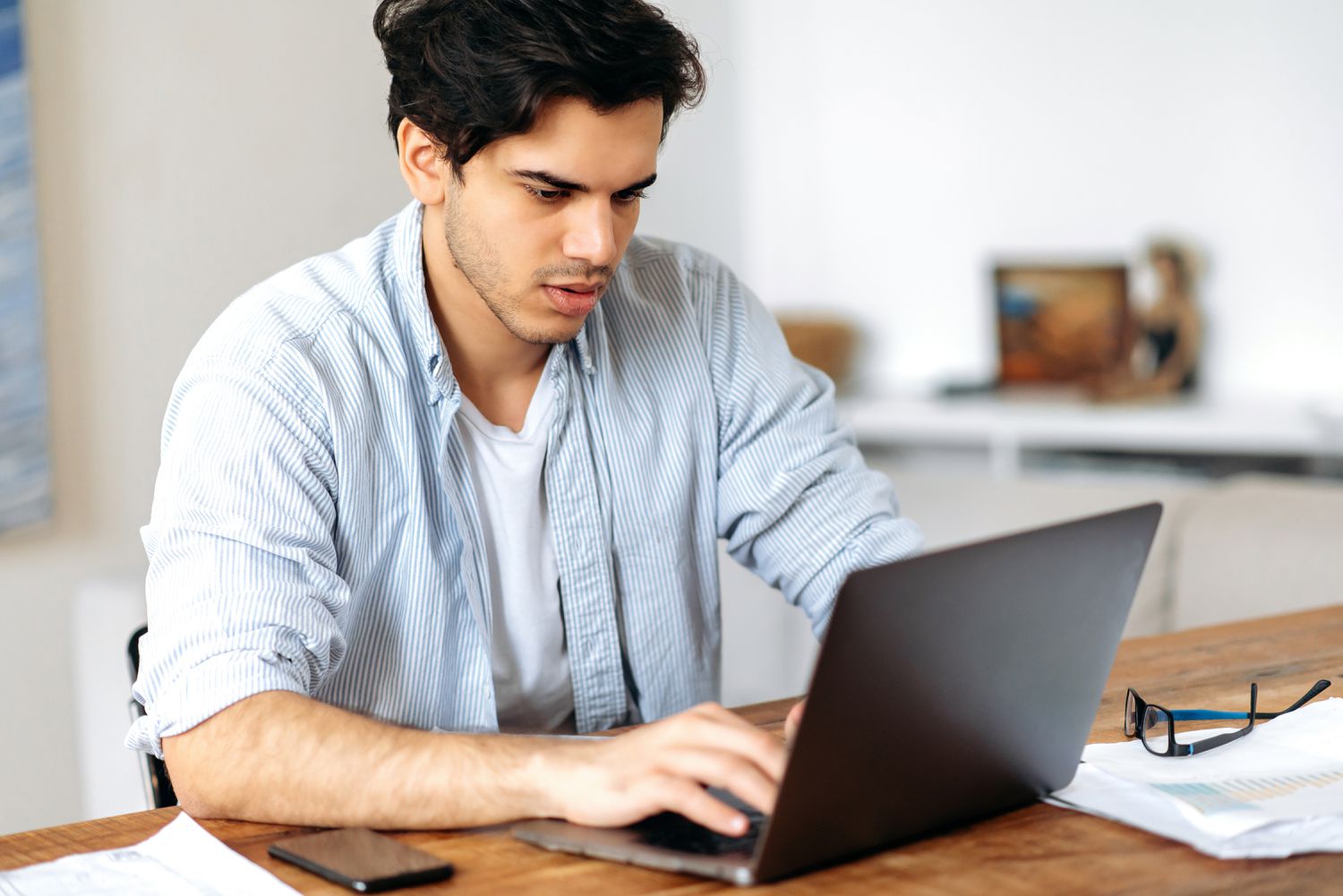 Young man sitting at his home desk and looking seriously at financial information on a laptop