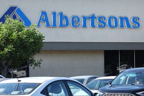 The tops of cars are seen in front of the side of an Albertsons grocery store, with a tree on the left side of the picture.
