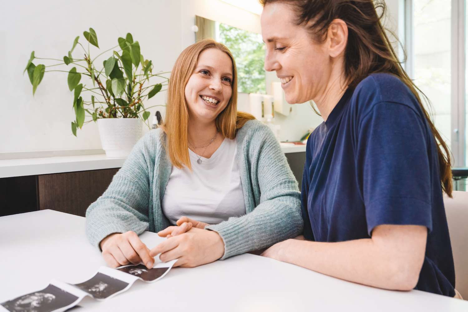 A joyful caucasian lesbian couple, holding hands while sitting at the gynecologist's desk, with their baby's ultrasound visible on the table.