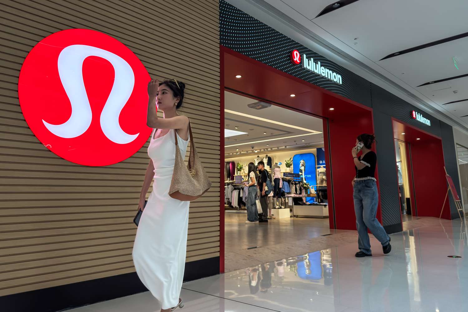  A woman in a white dress walks past a Lululemon store with its signature red logo displayed prominently on the wall, as shoppers browse inside the store