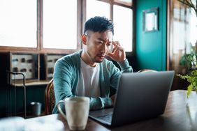 Worried young Asian man with his hand on head, using laptop computer at home, looking concerned and stressed out