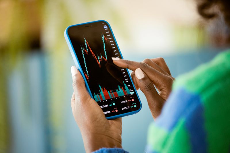 A close-up of a woman's hand, holding a cell phone and pointing to a stock chart on the screen. 