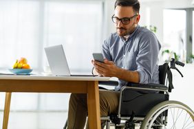 Man in a wheelchair in front of a desk with a laptop on it, looking at his phone.