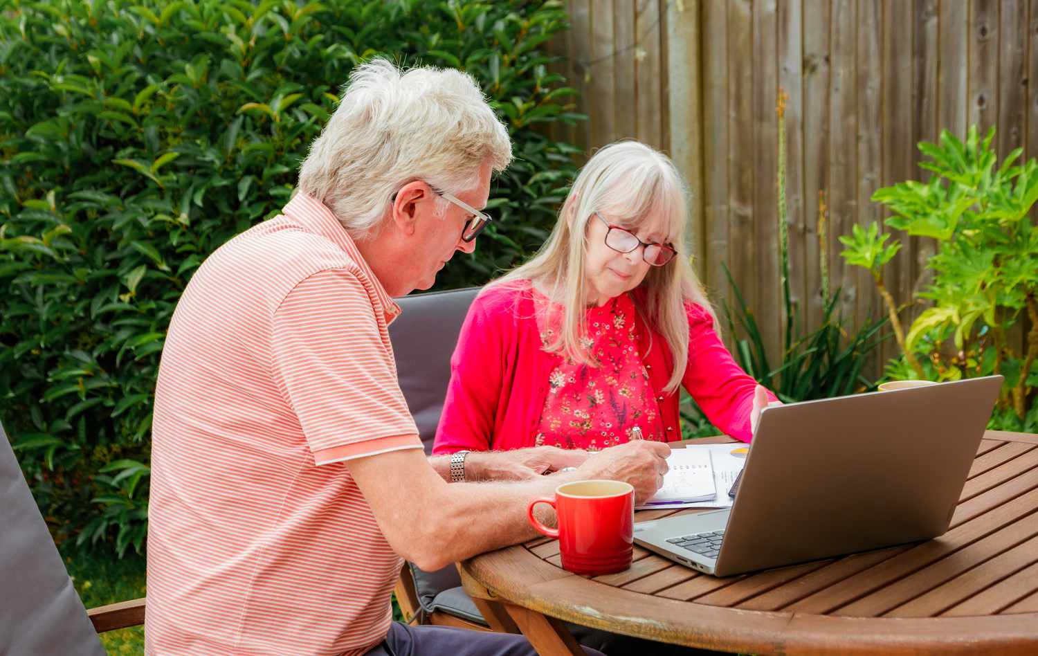 A couple checking their finances in the backyard with documents, a laptop, and coffee.