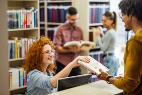 Happy female librarian taking a book from a male student in the library.