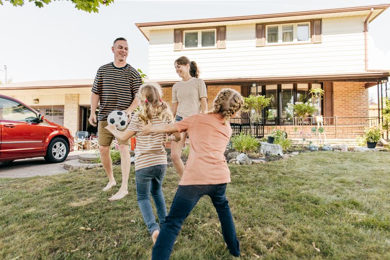 A family playing soccer in front of their house with a red car park in their driveway.