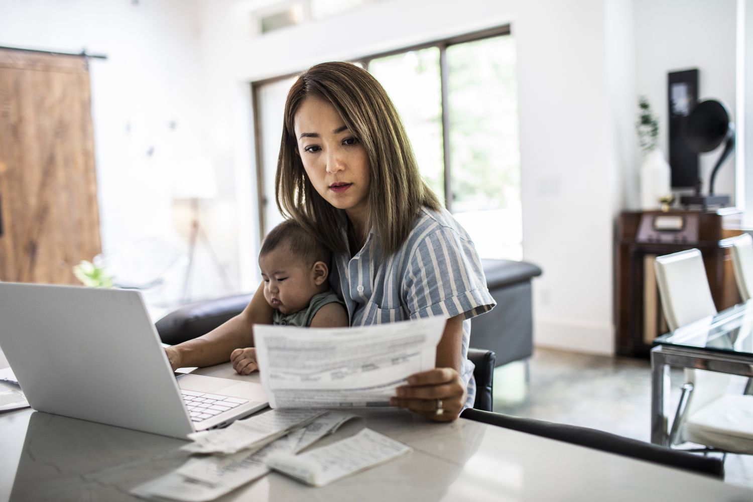 A woman sits at a table with a baby in her lap looking at paperwork and a laptop