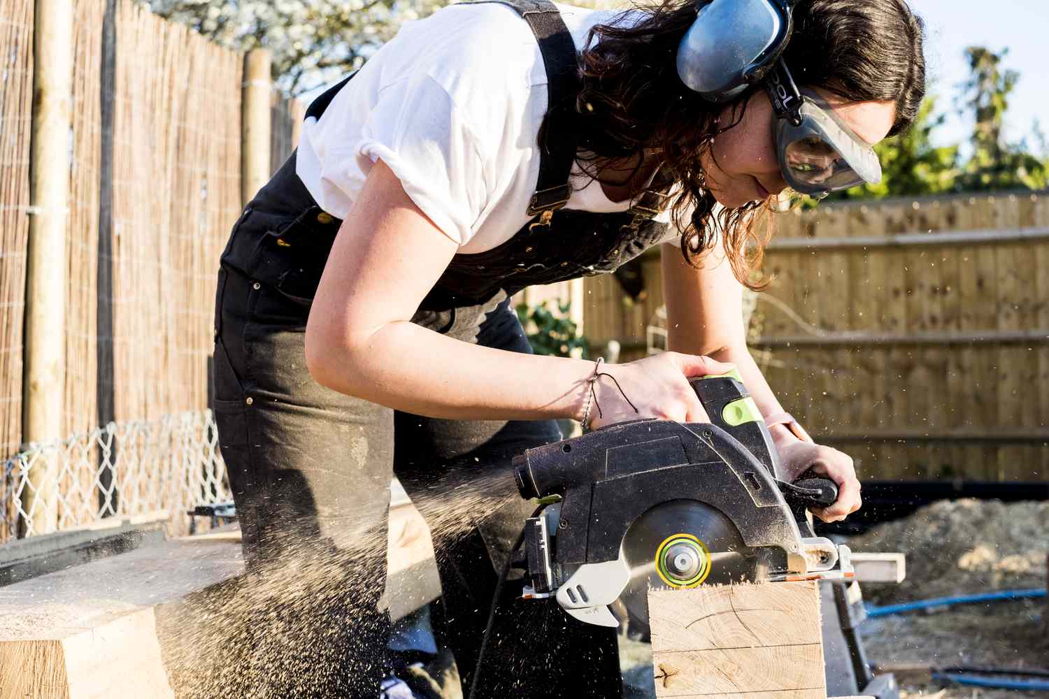 A person wearing protective goggles and ear protectors uses a circular saw to cut a piece of wood