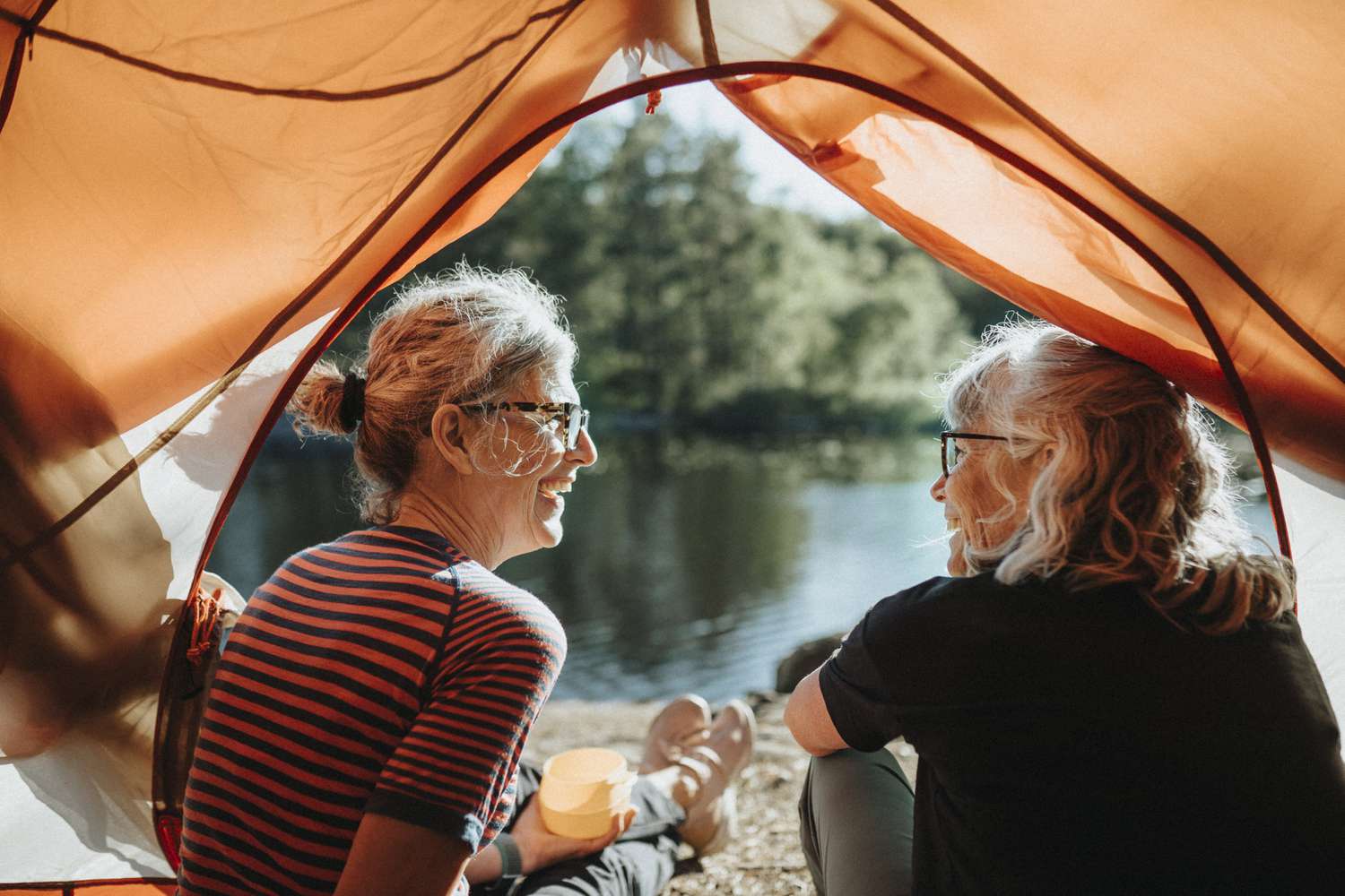 Two older women under a tent near the water at a campsite