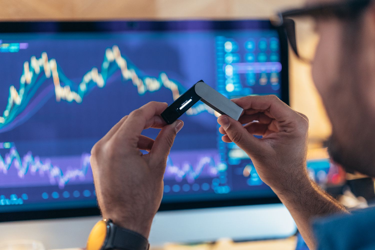 A man holds a cryptocurrency wallet while sitting in front of a computer screen displaying financial charts.