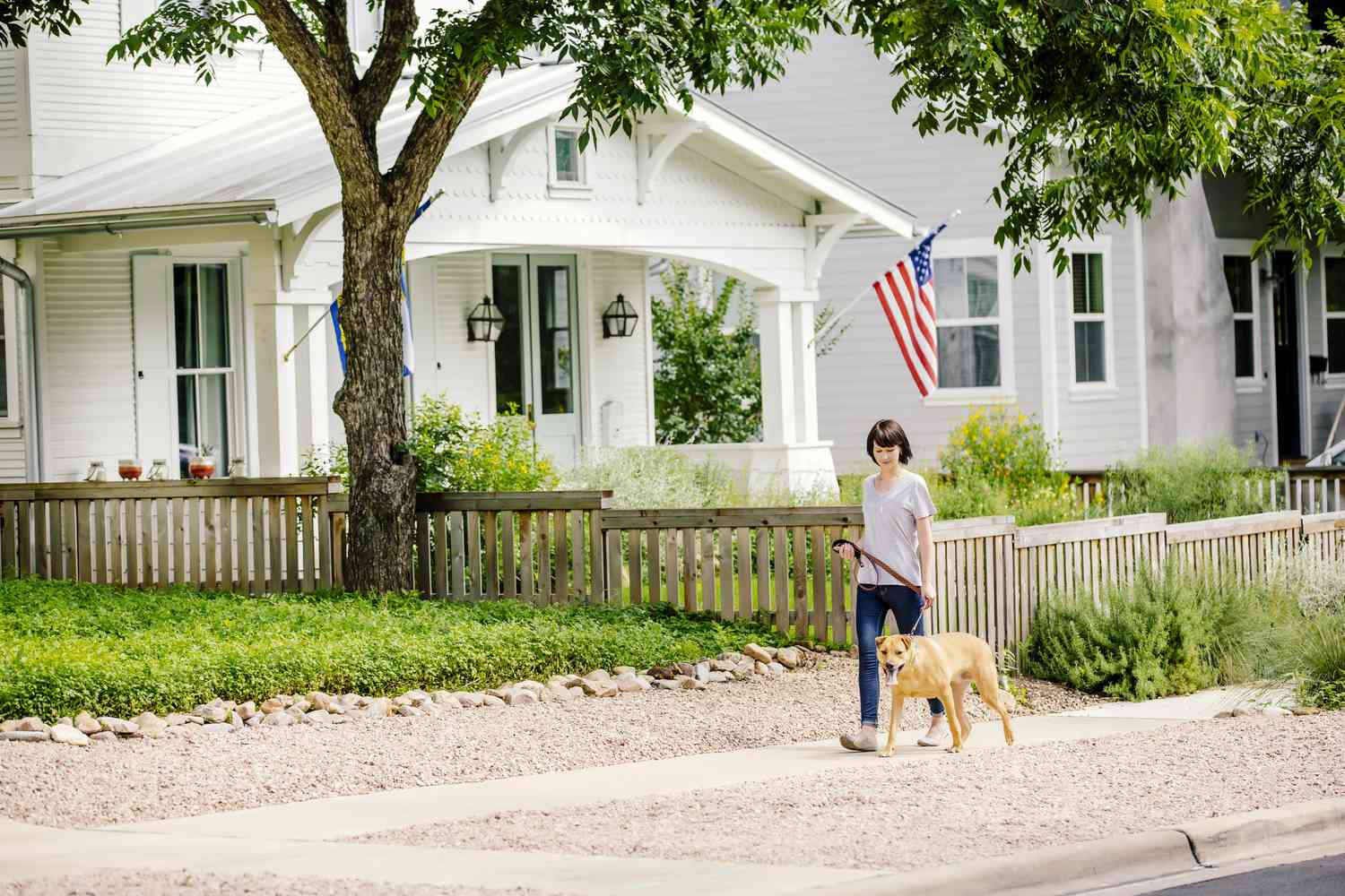 A woman walks her dog down the street in front of a white house with an American flag